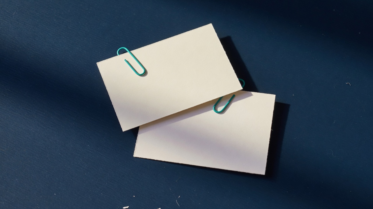 Two blank cream cards overlapping on a navy desk, each held by a teal paperclip
