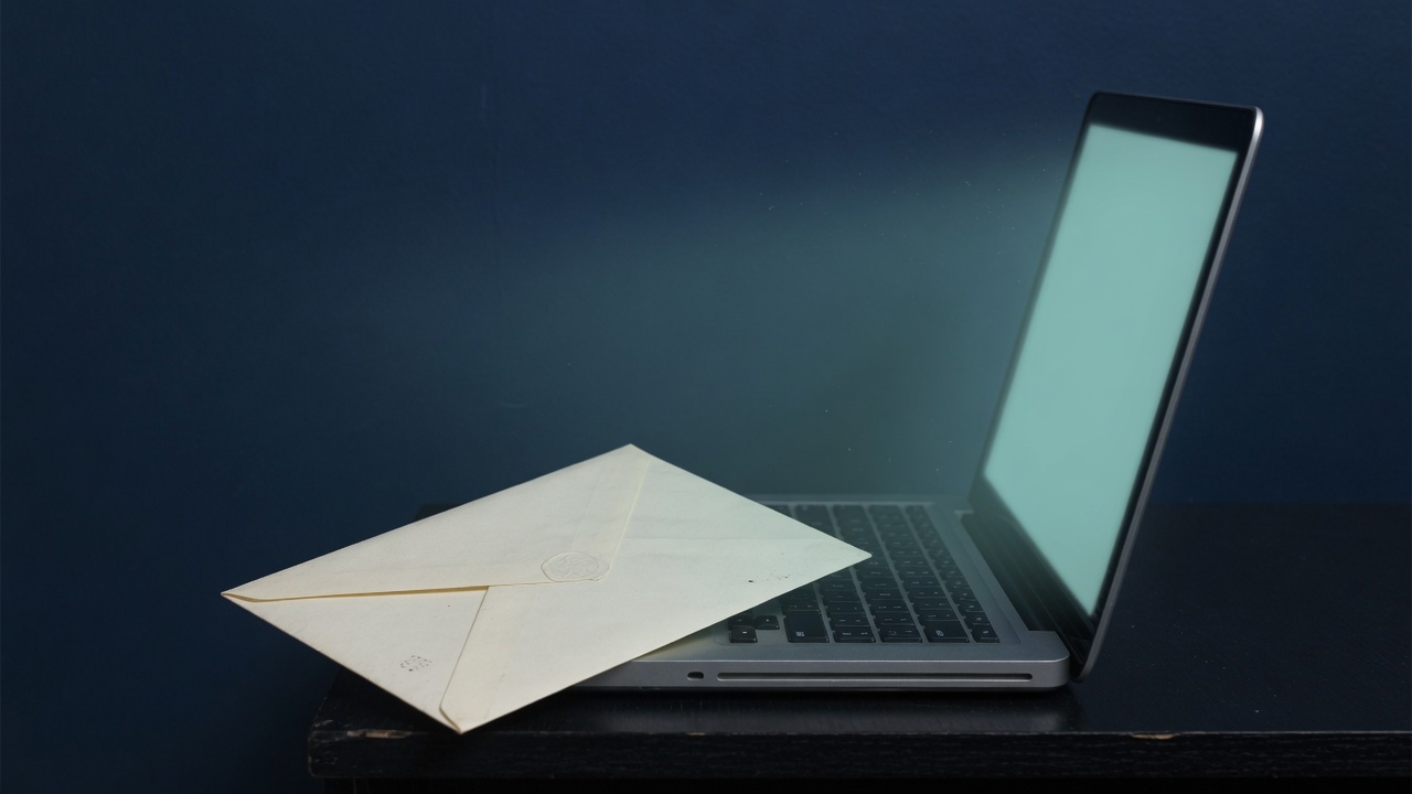 A laptop glowing teal on a dark navy desk beside one cream envelope