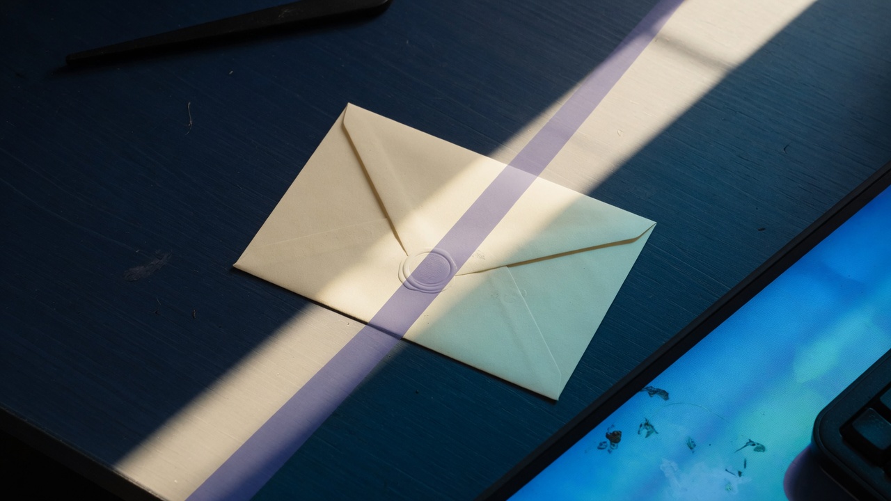 A dark navy desk split between daylight and a monitor glow, with one cream envelope in the middle