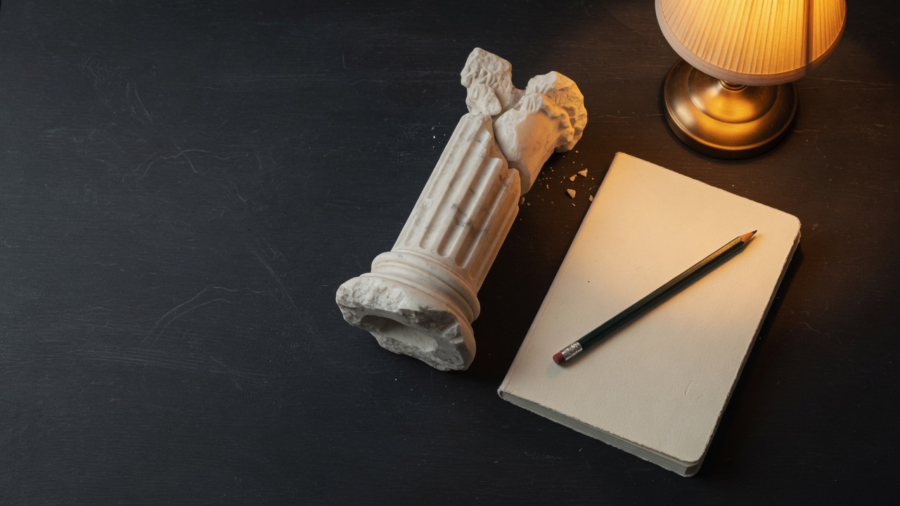 A broken white plaster column, a cream notebook, and a green pencil on a dark desk beside a lit lamp